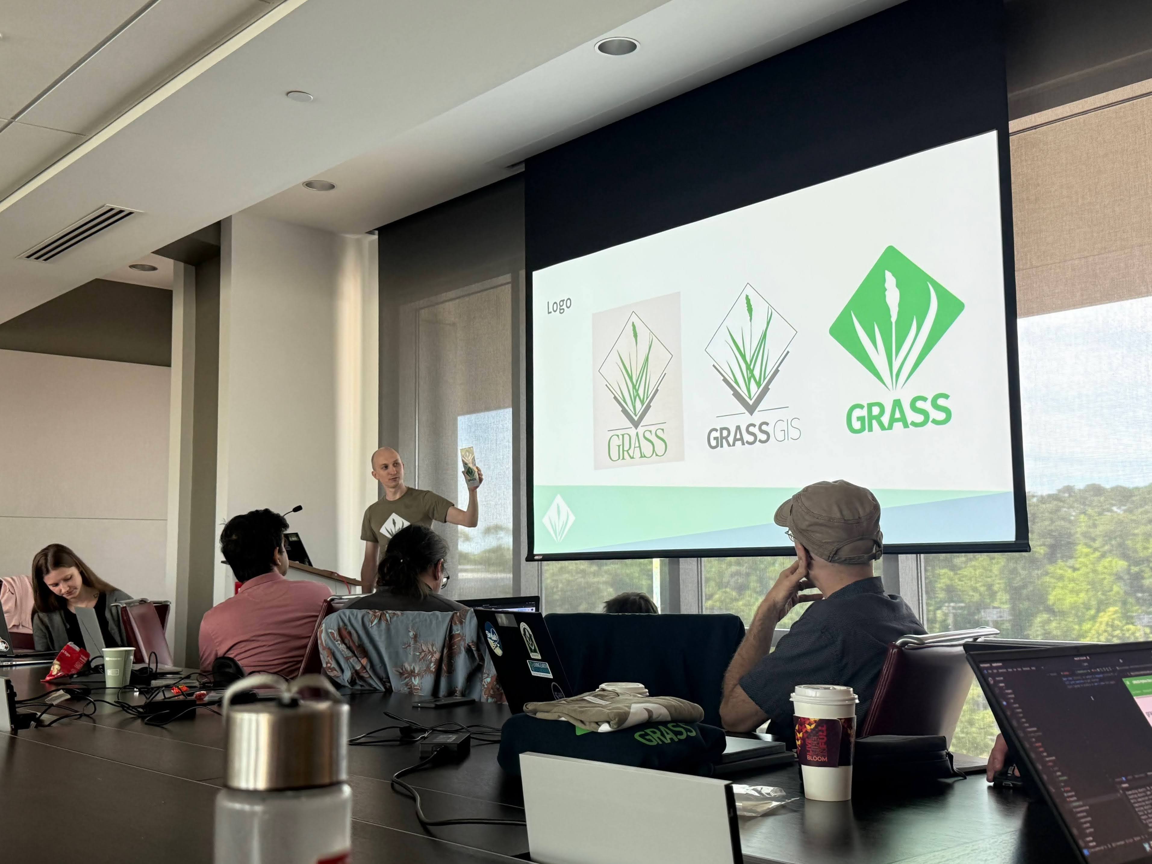 Person with a projector screen showing three versions of the GRASS logo (original, GIS version, and the new one)