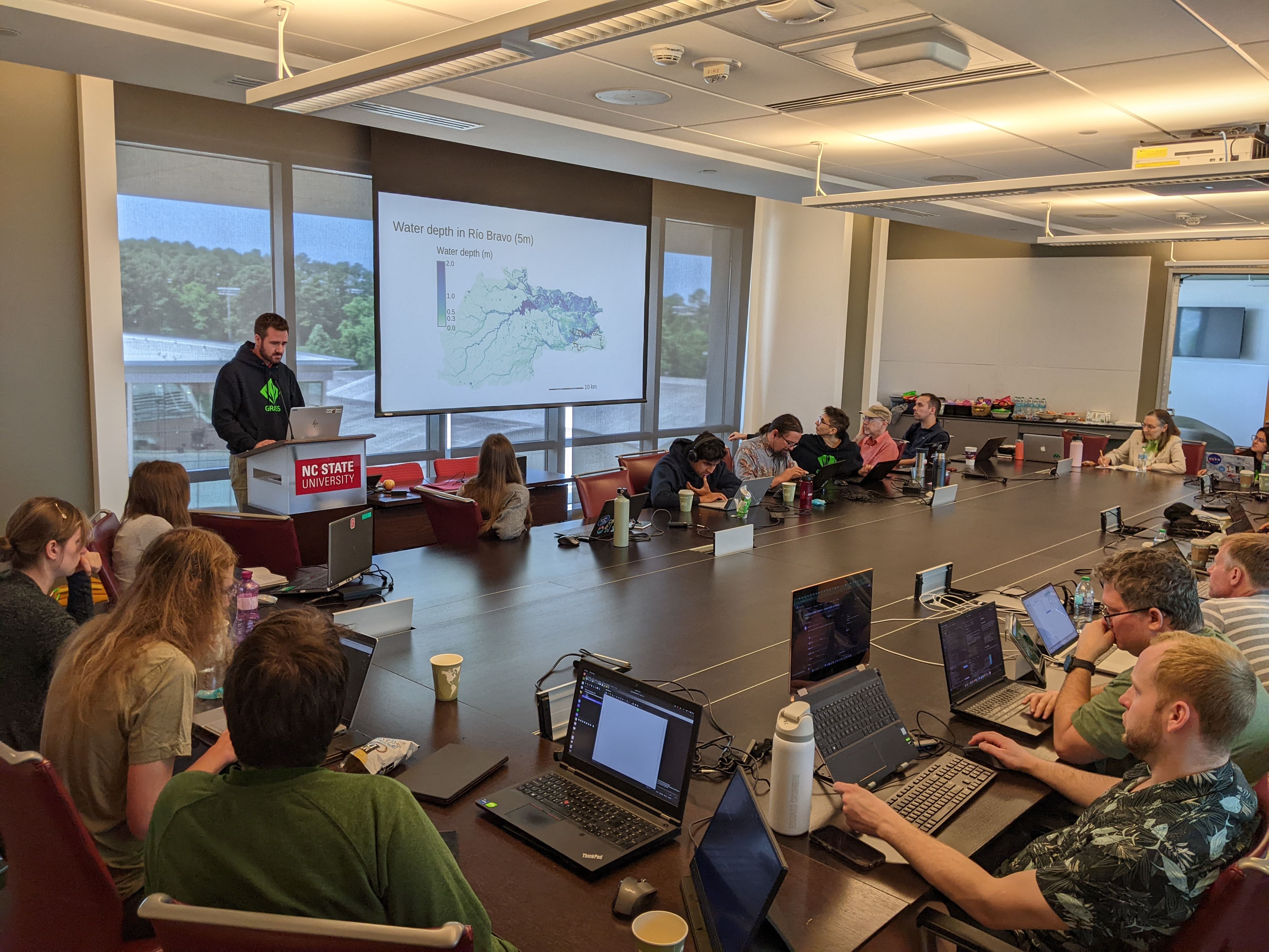 A person with slideshow in front of a room of people around a large conference table